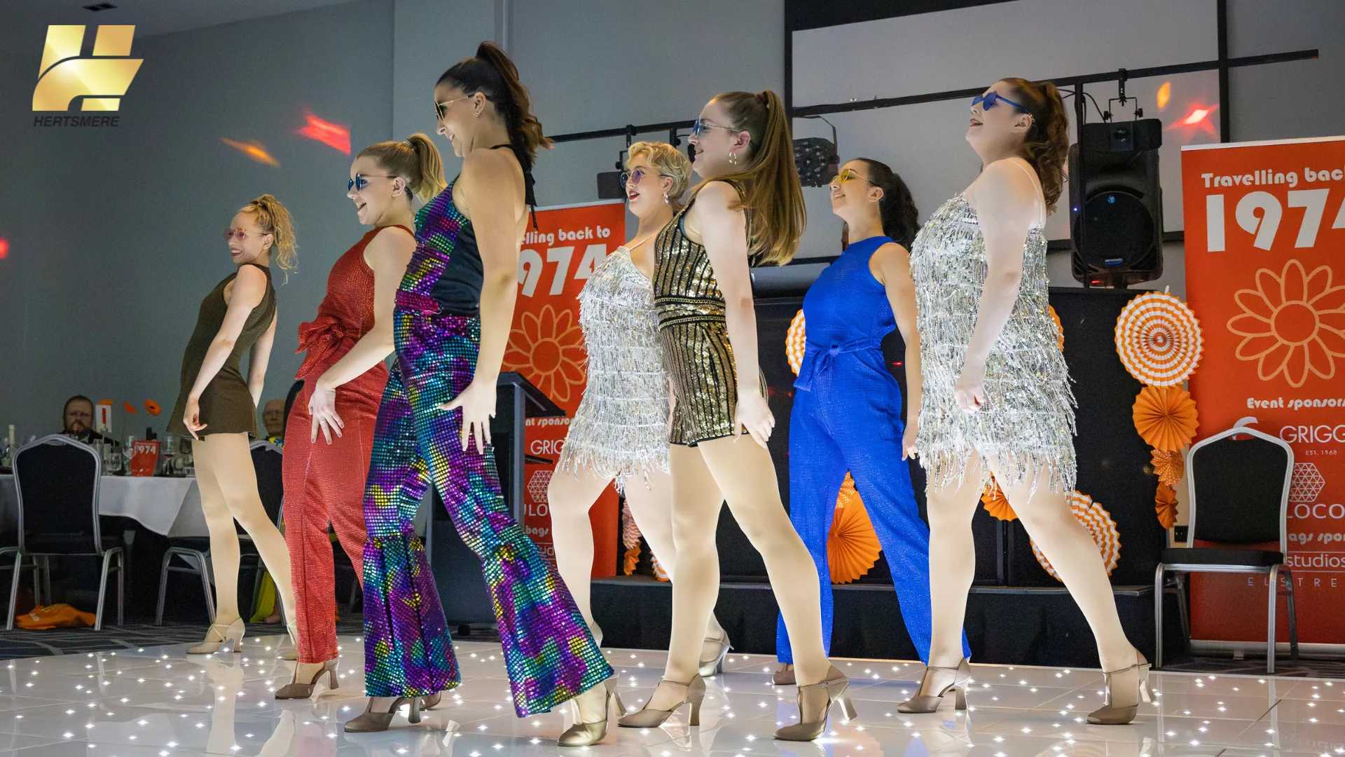 Students perform and dance in sync to a 1970s musical medley on glitter dancefloor.