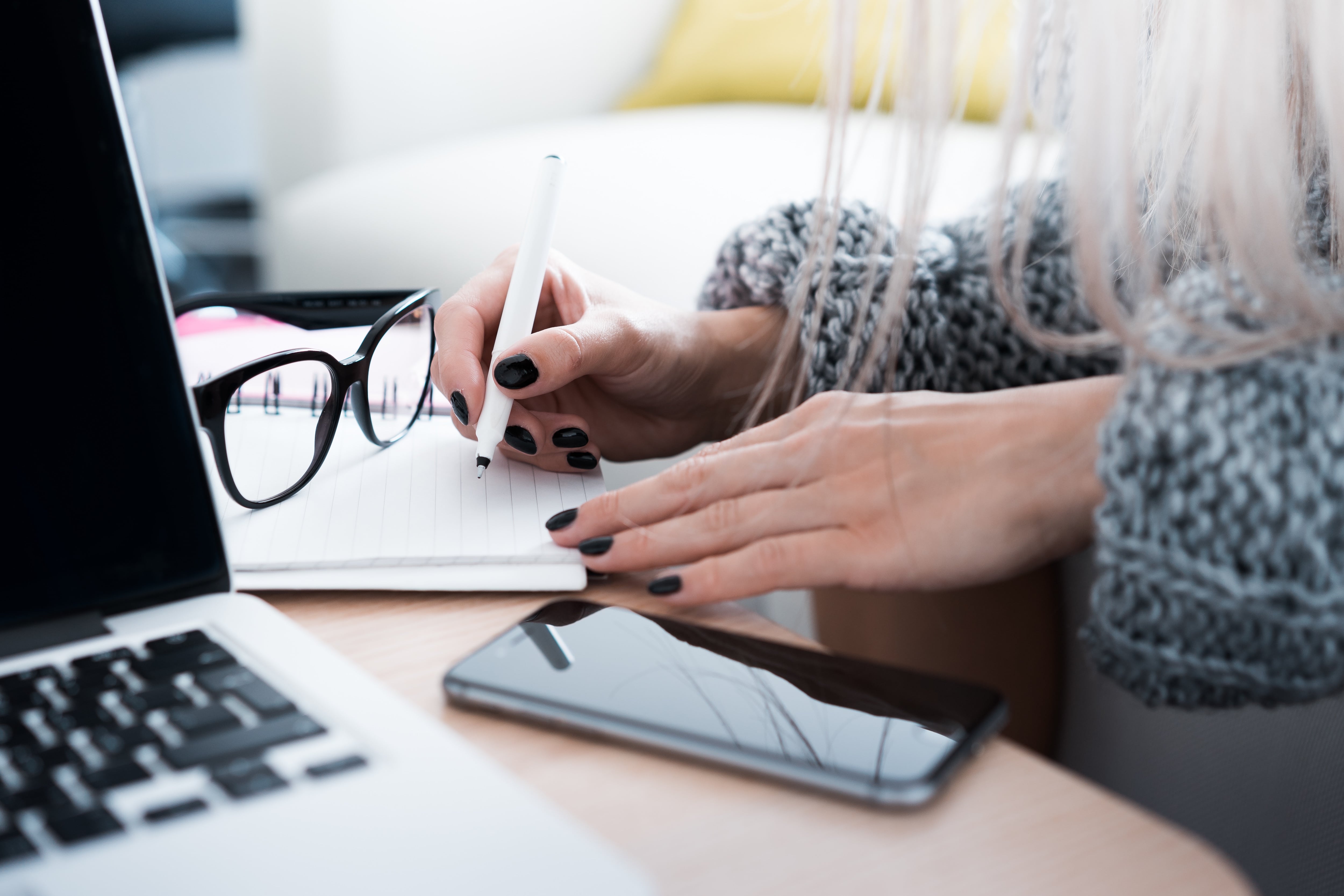 Woman writing notes at a desk with her mobile phone, glasses and computer nearby.