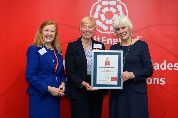 Chairwoman Audrey Adams and Trustee Margaret Craig-Gray receive Hidden Gem Accolade from Visit England employee in front of a red background.